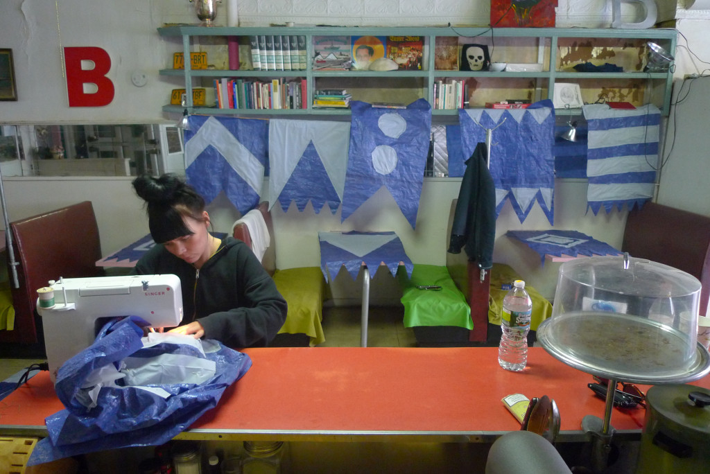 Milja Havas sewing the flags from tarpaulin. Finished flags hanging at the background. At the Sunview Luncheonette, Greenpoint. (photo by Johannes Rantapuska, 2014)